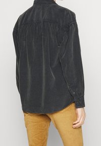 Persona che indossa una camicia nera in velluto a coste a maniche lunghe e pantaloni in velluto a coste giallo senape, in piedi con la schiena rivolta verso la fotocamera.