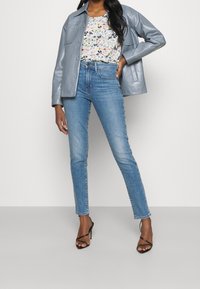 Giubbotto di pelle azzurro chiaro sopra una maglietta floreale, abbinato a jeans azzurro chiaro aderenti e décolleté neri con cinturini. Texture liscia, vestibilità sartoriale.