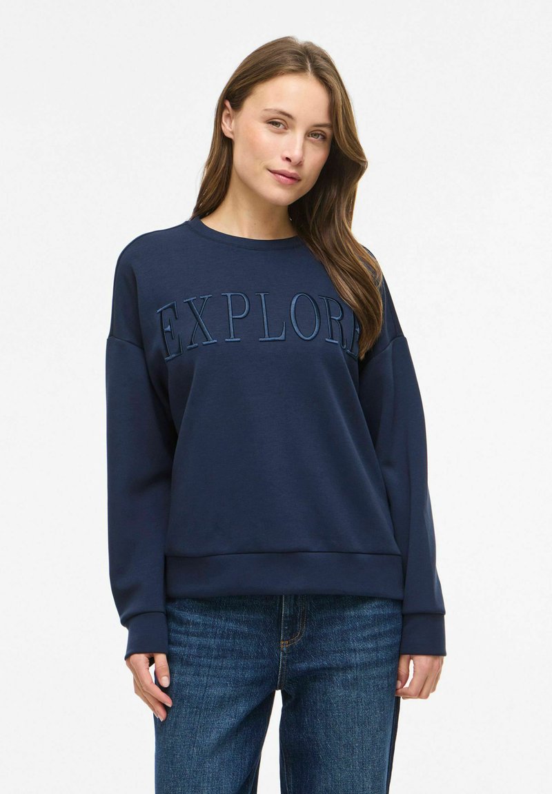 Femme aux longs cheveux bruns portant un sweat-shirt bleu marine avec l'inscription "EXPLORE" et un jean bleu, debout devant un fond blanc uni.