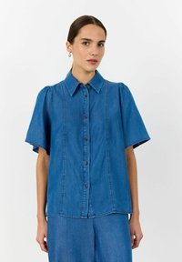 Niebieska koszula jeansowa z zapięciem na guziki z przodu, krótkimi bufiastymi rękawami, klasycznym kołnierzykiem oraz pionowymi szwami.