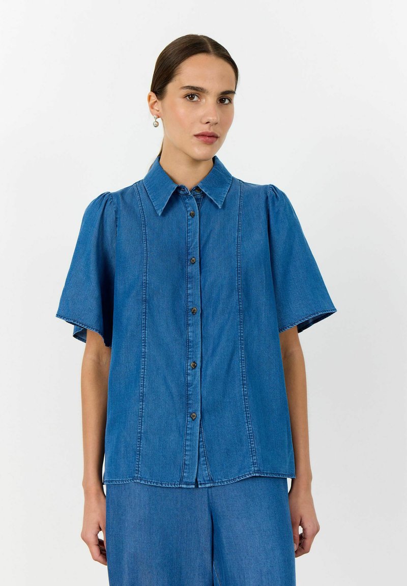 Niebieska koszula jeansowa z zapięciem na guziki z przodu, krótkimi bufiastymi rękawami, klasycznym kołnierzykiem oraz pionowymi szwami.