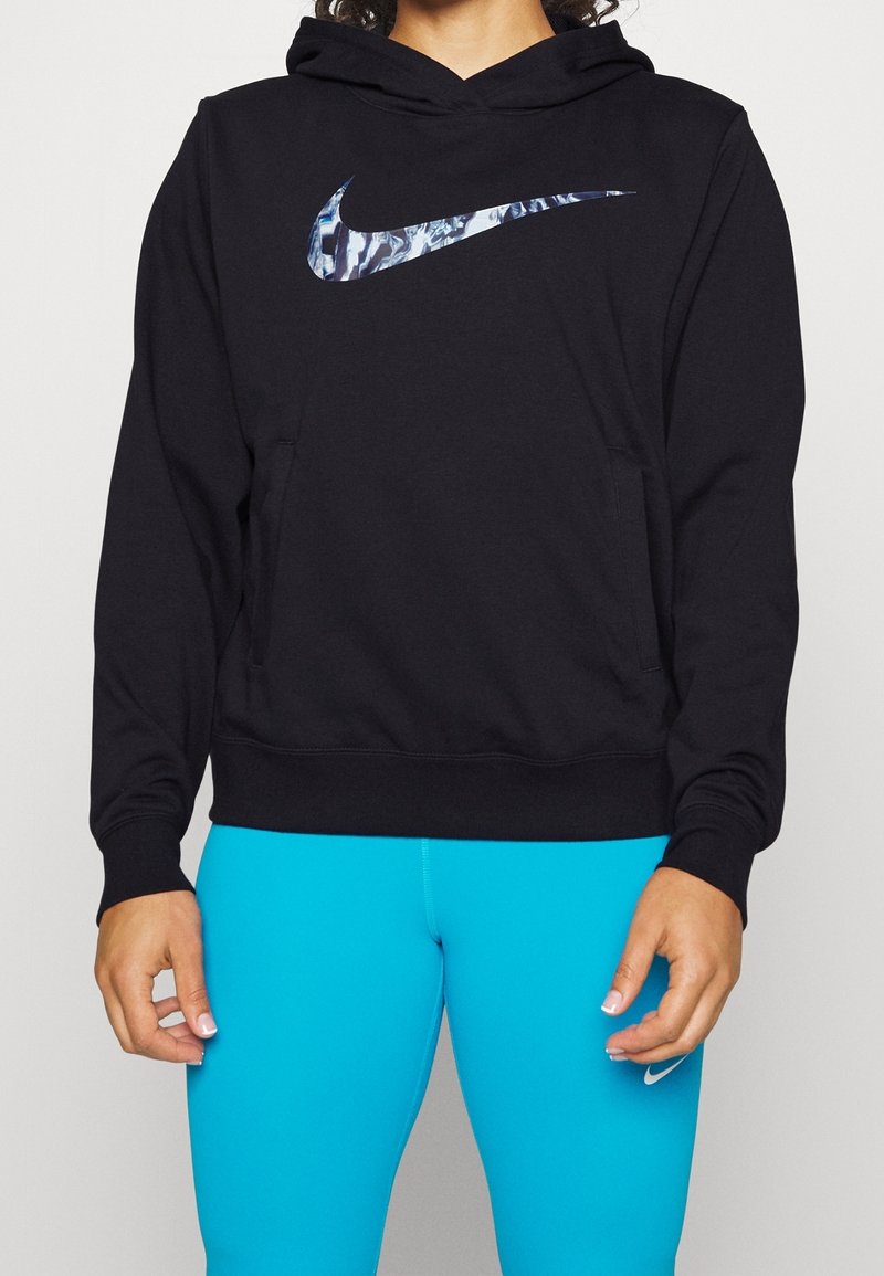 Person in einem schwarzen Hoodie mit einem blauen, gemusterten Nike-Logo und hellblauen Nike-Leggings, der vor einem schlichten Hintergrund steht.