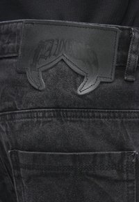 Melni džinsa bikses ar fakturētu audumu. Nozīmīgas detaļas ir liela melna ādas plāksteris ar reljefu dizainu jostasvietā.