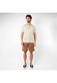 Chemise à manches courtes de couleur jaune pâle avec un motif floral bleu, assortie à un short marron et des baskets noires. Dotée d'un col standard et de détails de boutons.