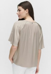 Beige blus med kort ärm i mjukt tyg, med en plisserad bak och en knappstängning vid halsen. Enkel, avslappnad passform.
