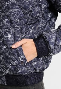 Marineblaue Steppjacke mit einem weißen geometrischen Muster, elastischen Bündchen und einer Seitentasche. Die gesteppten Texturen bieten Isolierung und Wärme.