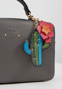Sac à main gris LYDC London avec poignée noire et breloque colorée en forme de cactus décorée de fleurs rouges et orange.