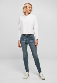 Camisa de polo blanca de manga larga y corte corto con cuello, combinada con jeans ajustados azules y zapatillas blancas con detalles verdes.