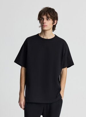 Giovane uomo con capelli castani arruffati che indossa una maglietta nera a maniche corte e pantaloni neri, guarda di lato.