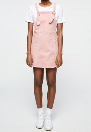Osoba ubrana w różową sukienkę bez rękawów z przednimi kieszeniami na białej koszulce, białe skarpety oraz białe trampki stojąca na jednolitym tle.