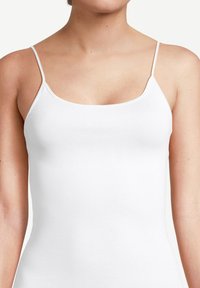Donna che indossa un top attillato bianco a spalline sottili, che mostra le spalle, le braccia superiori e la parte superiore del petto, contro uno sfondo neutro.