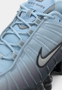 Nike Shox tenisice u svijetlo plavoj mrežici s sivim naglascima, koje imaju karakterističan dizajn potplata s cilindričnim amortizerima i teksturiranim površinama.