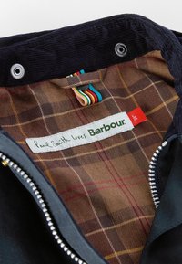 Jaqueta de tecido exterior preto com forro xadrez castanho. Apresenta um fecho metálico e uma etiqueta de marca que diz "Paul Smith ama Barbour."