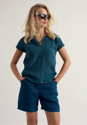 Mujer con camiseta de manga corta texturizada color verde azulado oscuro, pantalones cortos a juego, gafas de sol negras y collar turquesa, con las manos en los bolsillos, mirando hacia un lado.