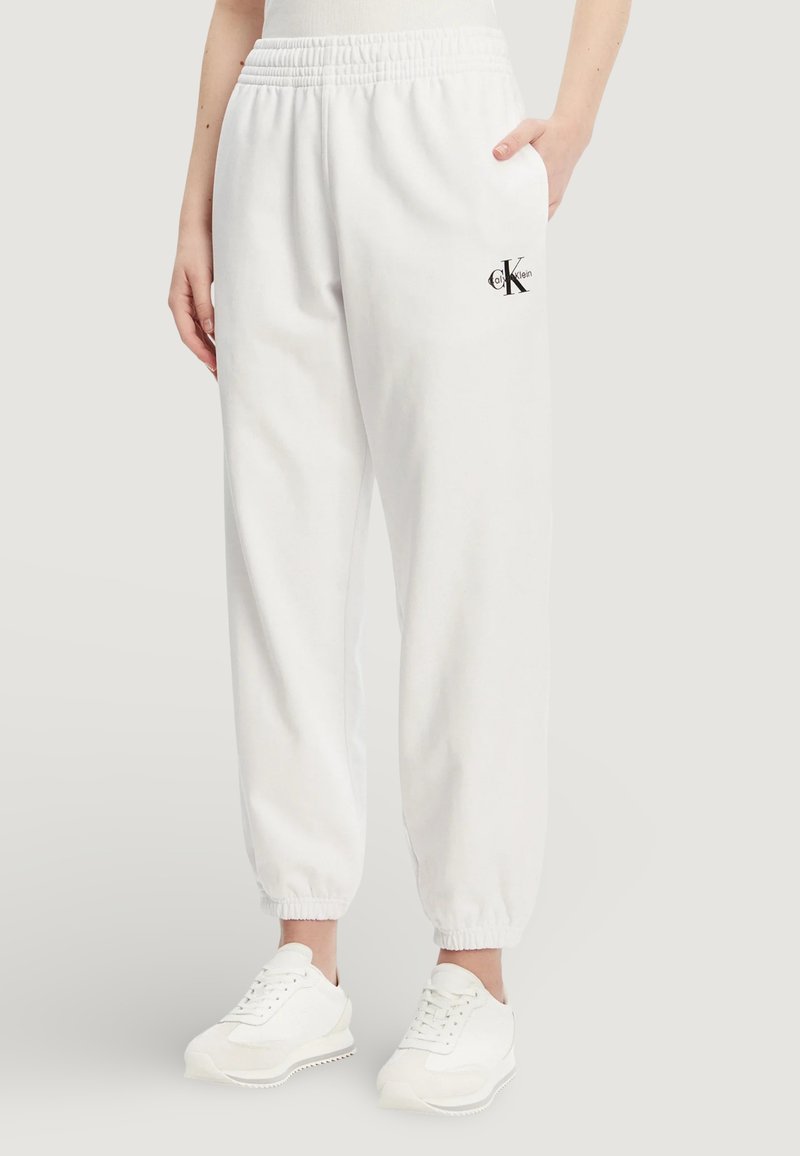 Bílé bavlněné tepláky s elastickým pasem, manžetami na kotnících, bočními kapsami a malým černým logem na levém stehně. Nošeny s bílými teniskami.