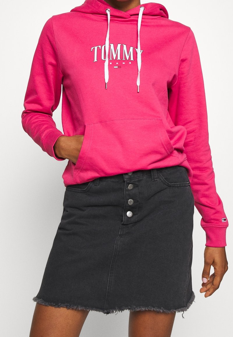 Sudadera fucsia con texto blanco "TOMMY", con un bolsillo frontal y cordones; combinada con una falda de mezclilla negra con dobladillo deshilachado y botones.