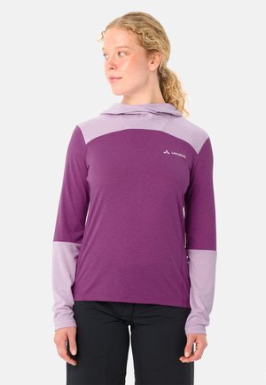 Vrouw draagt een paars en licht lavendel gekleurde lange mouwen hoodie, staand en licht naar links kijkend tegen een effen achtergrond.