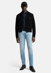 Giacca nera in suede con tasche frontali, camicia blu a bottoni, jeans azzurri, e stivaletti neri. Indossato con un cappellino nero.
