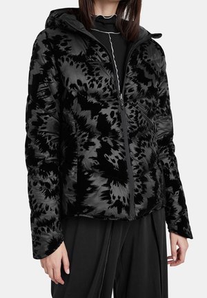 Femme portant une doudoune à capuche noire et gris foncé à motifs, un haut noir et un pantalon noir ample.