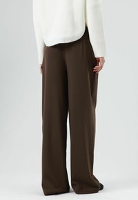 Bruine wijde broek met een gladde textuur, hoge taille en relaxte pasvorm. Gecombineerd met een wit gebreid topje, gezien vanaf de achterkant.