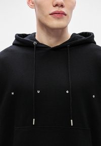 Sudadera negra de tela suave con un bolsillo frontal, cordones ajustables y el logotipo "HUGO" en letras blancas en el pecho.