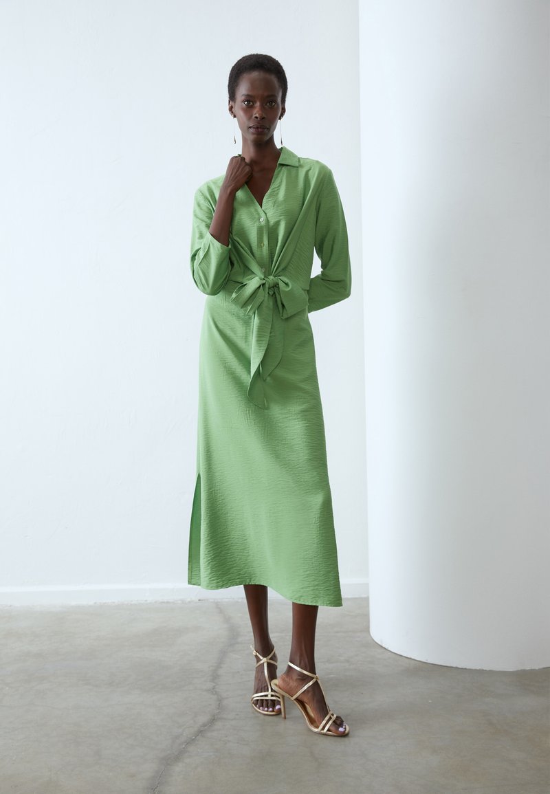 Knie-lange jurk met lange mouwen in textuurstof in lichtgroen, met een striktaille en een zijsplit. Gecombineerd met metallic hakken met bandjes.
