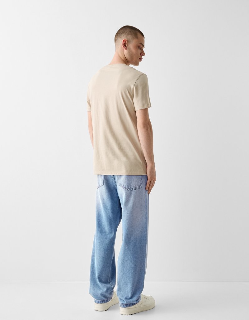 Beige Baumwoll-T-Shirt, kurzärmlig, mit Rundhalsausschnitt. Getragen mit hellblauen, ausgewaschenen, weiten Jeans und weißen Sneakers. Lässiger Schnitt.