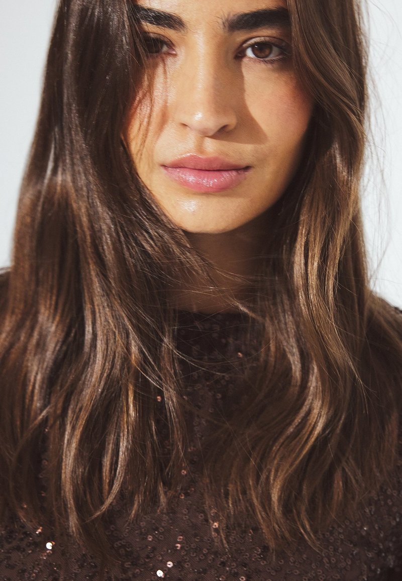 Close-up van een persoon met lang, golvend bruin haar, gekleed in een donkere, met pailletten bedekte outfit, met zichtbare huidtextuur en natuurlijke lipkleur.