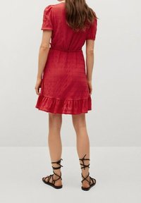Rotes knielanges Kleid mit kurzen Ärmeln, strukturiertem Stoff, tailliertem Schnitt und Rüsche am Saum. Kombiniert mit schwarzen Sandalen mit Riemen.