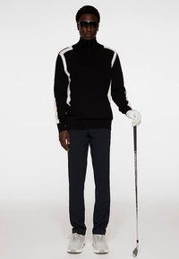 Svart zip-up tröja med vita sidodetaljer, svarta byxor och gråa sneakers. Personen håller i en golfklubba, bär solglasögon och en golfhandske.