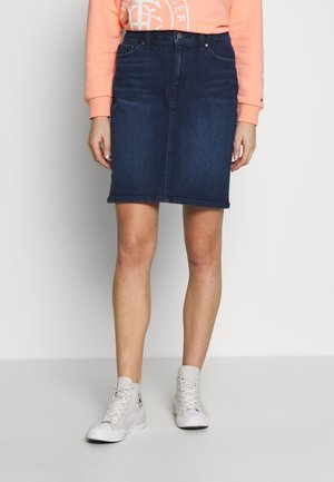 Femme portant une jupe en denim bleu foncé jusqu'aux genoux, un sweat pêche et des baskets blanches montantes, debout devant un fond uni.