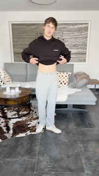 Black hoodie, gray sweatpants, and white sneakers. Sitting room background with a gray couch, round table, and patterned rug.