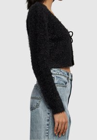 Pull noir, court et flou, avec de longues manches et un détail à lacets sur le devant, porté avec un jean en denim bleu clair taille haute.