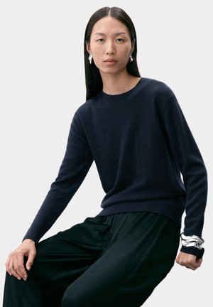 Navy blue knitted sweater with long sleeves and round neckline, paired with loose black trousers. Silver modern cuff bracelet on wrist.