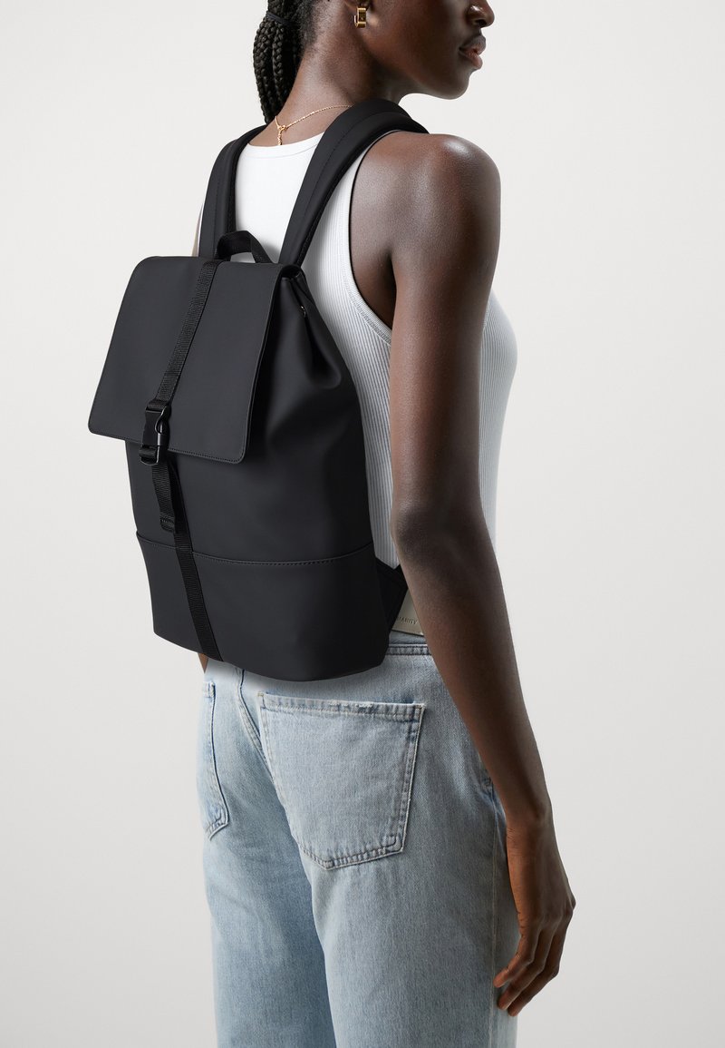 Black backpack made of smooth material; features a flap closure, adjustable straps, and a flat shape. Worn on a person against a neutral background.