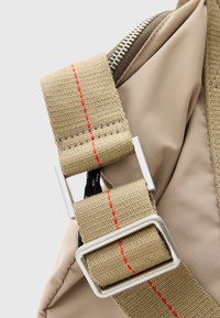 Correa de mochila caqui con costuras rojas, herrajes plateados y textura suave. La abertura con cremallera revela un forro beige. Diseño duradero y funcional.