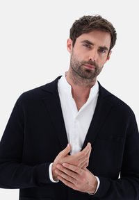 Marinefarbener Cardigan mit modernem Design, über einem weißen Hemd getragen. Der Stoff wirkt strukturiert und hat eine Taschenakzentuierung auf der linken Seite.