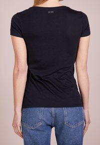 Černé bavlněné tričko s kulatým výstřihem a krátkými rukávy, na zadní straně s malým logem. V kombinaci s vysokými modrými džínami.