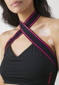 Haut de maillot de bain noir avec un design en croisement et des bretelles d'accentuation noires, rouges et bleues. Comprend le logo Speedo en rouge sur le côté.