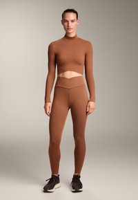 Bruine lange mouwen crop top met een uitsnede en bijpassende hoge taille leggings, beiden gemaakt van een gladde, elastische stof. Zwarte sportschoenen.