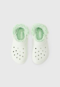 Weiße Clogs mit Belüftungslöchern und einem hellgrünen Fleece-Innenfutter entlang der Öffnung, von oben auf einem weißen Hintergrund gezeigt.
