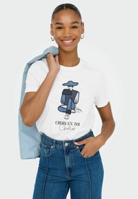 T-shirt in cotone bianco con una grafica di una figura seduta in blu, con il testo "Crois en toi, Chérie." Abbinato a jeans in denim a vita alta.
