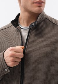 Mężczyzna zapinający brązową, teksturowaną kurtkę na białą koszulę z czarno-szarym kołnierzykiem, widoczny od podbródka do klatki piersiowej.