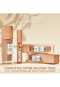 Quatre produits de maquillage de fondation dans des bouteilles en verre rectangulaires avec des bouchons en bronze. Les étiquettes comprennent des accents verts et des détails sur les ingrédients.