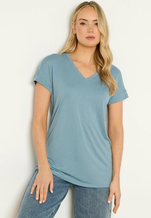 Frau mit langen blonden Haaren, die ein hellblaues T-Shirt mit V-Ausschnitt und kurzen Ärmeln sowie blaue Jeans trägt, steht vor einem schlichten hellen Hintergrund.