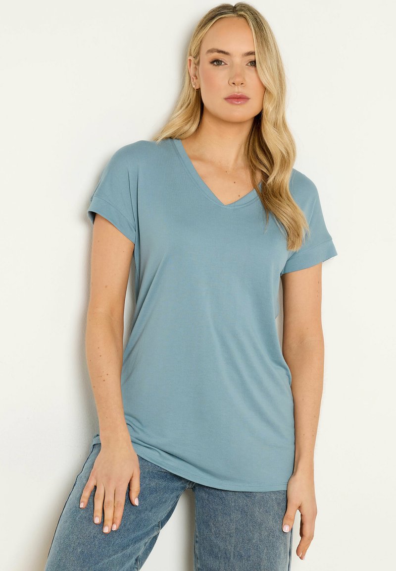 Mujer con cabello largo y rubio con una camiseta de manga corta azul claro con cuello en V y jeans azules, de pie contra un fondo claro y liso.