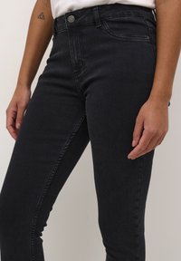 Jean skinny en denim délavé foncé, doté d'une taille haute, de poches avant et d'une fermeture à bouton en métal à la taille. Texture lisse.