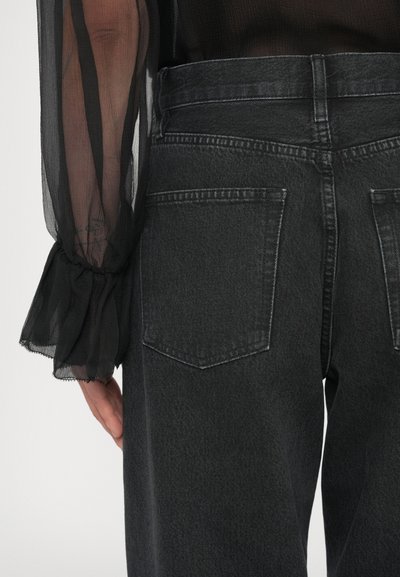 Calças de ganga cinza escuro com cintura alta, apresentando dois bolsos traseiros e uma textura suave. A peça é combinada com uma manga de blusa preta transparente.