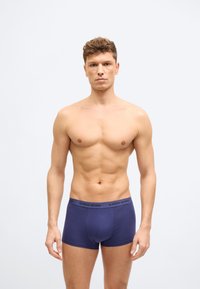 Modèle masculin athlétique aux cheveux bouclés portant un boxer Björn Borg bleu marine, debout devant un fond gris clair uni.