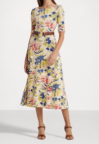 Blommig klänning med korta ärmar, prydd med rosa, blå och gröna blommor på en krämfärgad bakgrund. Inkluderar ett brunt bälte och en utställd kjoldesign.